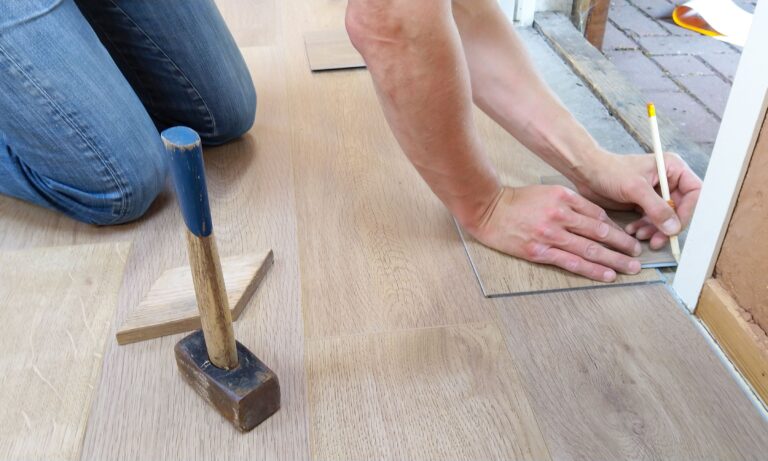 A person installs light-colored laminate flooring, using a pencil and rubber mallet to fit the planks.